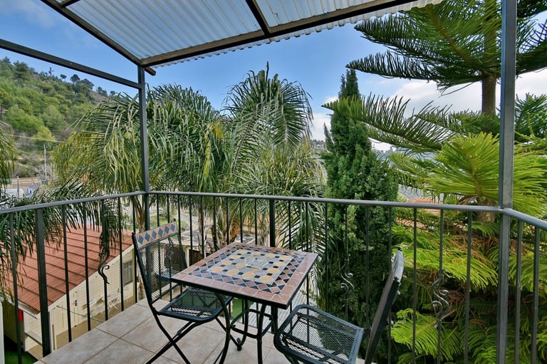 Outdoor deck with pergola, checkered table, chairs, and surrounding palm trees and green landscape