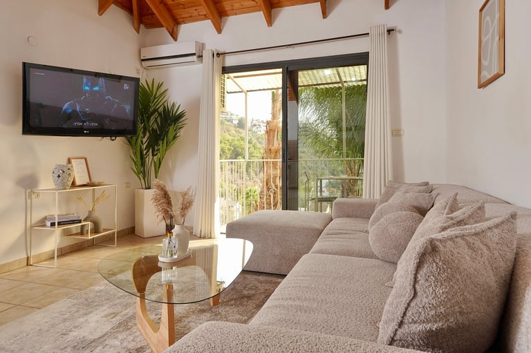 Modern living room with gray sectional sofa, round glass coffee table, wall-mounted TV, indoor plants, and sliding glass doors opening to a balcony
