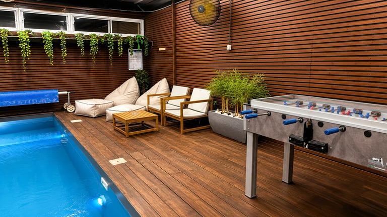 Indoor recreational facility with illuminated blue pool, wooden deck, lounge chairs, foosball table, and decorative plants under wood-paneled ceiling