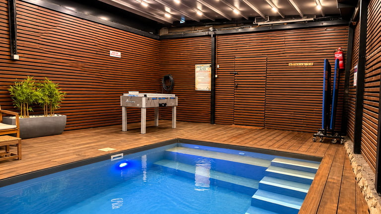 Indoor swimming pool with wooden deck and walls, stairs visible, exercise equipment, potted plant, and warm lighting